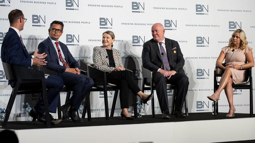 Five people wearing business attire, sitting on a stage in a row as part of a panel.